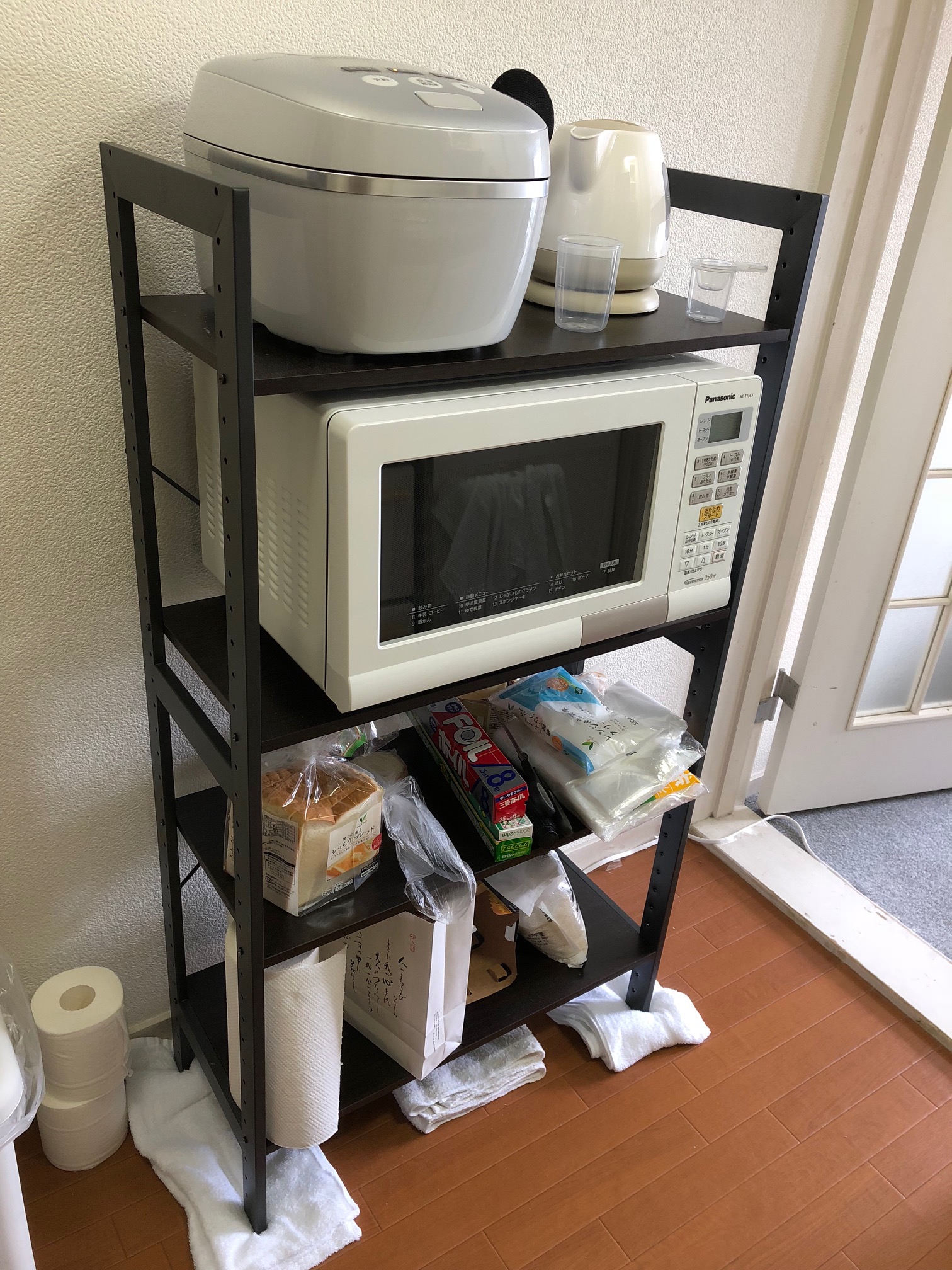 (Caption) I’m perhaps unreasonably proud for picking and assembling this shelf, but I just love how it looks and how perfectly it fits in there. From top to bottom, left to right, there’s rice cooker, kettle, microwave oven/toaster/regular oven all in one (not kidding) and then just some generic storage. The towels are unfortunately necessary for not damaging the incredibly fragile floor that looks like wood but is not wood at all.